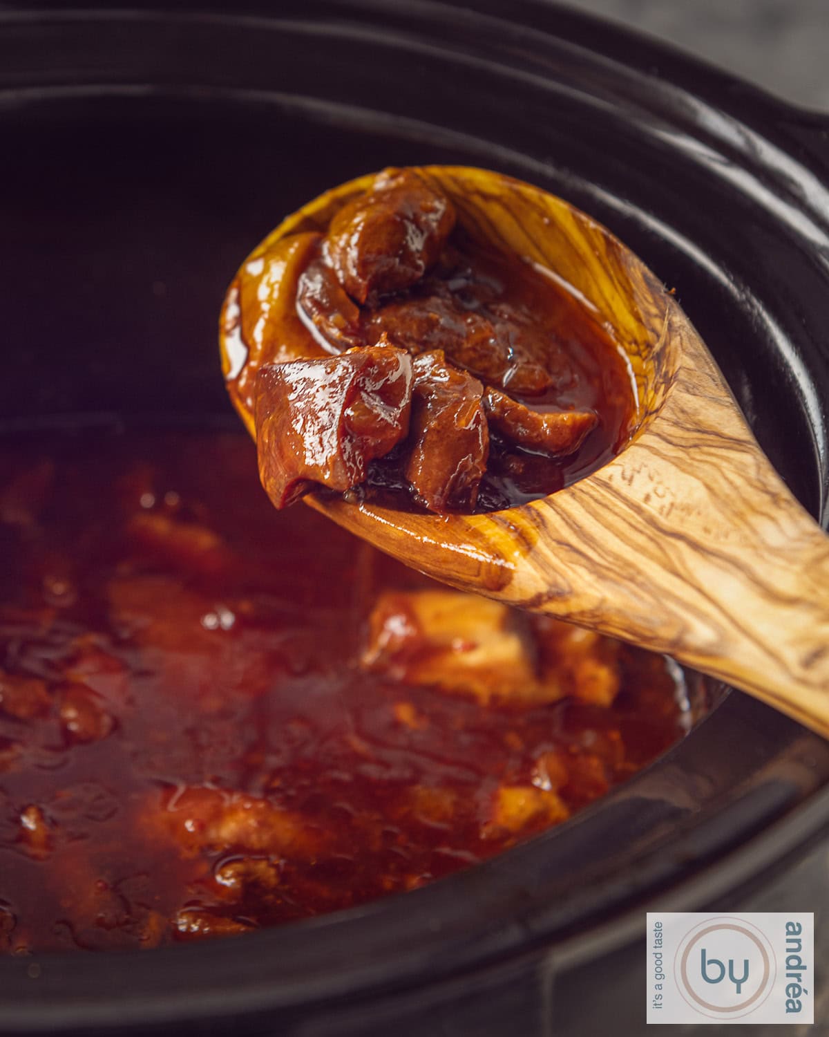 A slow cooker with a wooden spoon with chicken cubes on it. Ajam pangang is in the slow cooker