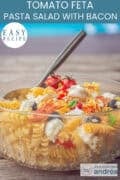 Part of a glass dish with olives, feta, roasted tomatoes, pasta, bacon and basil. A text on top: Tomato Feta Pasta Salad with Bacon, easy recipe