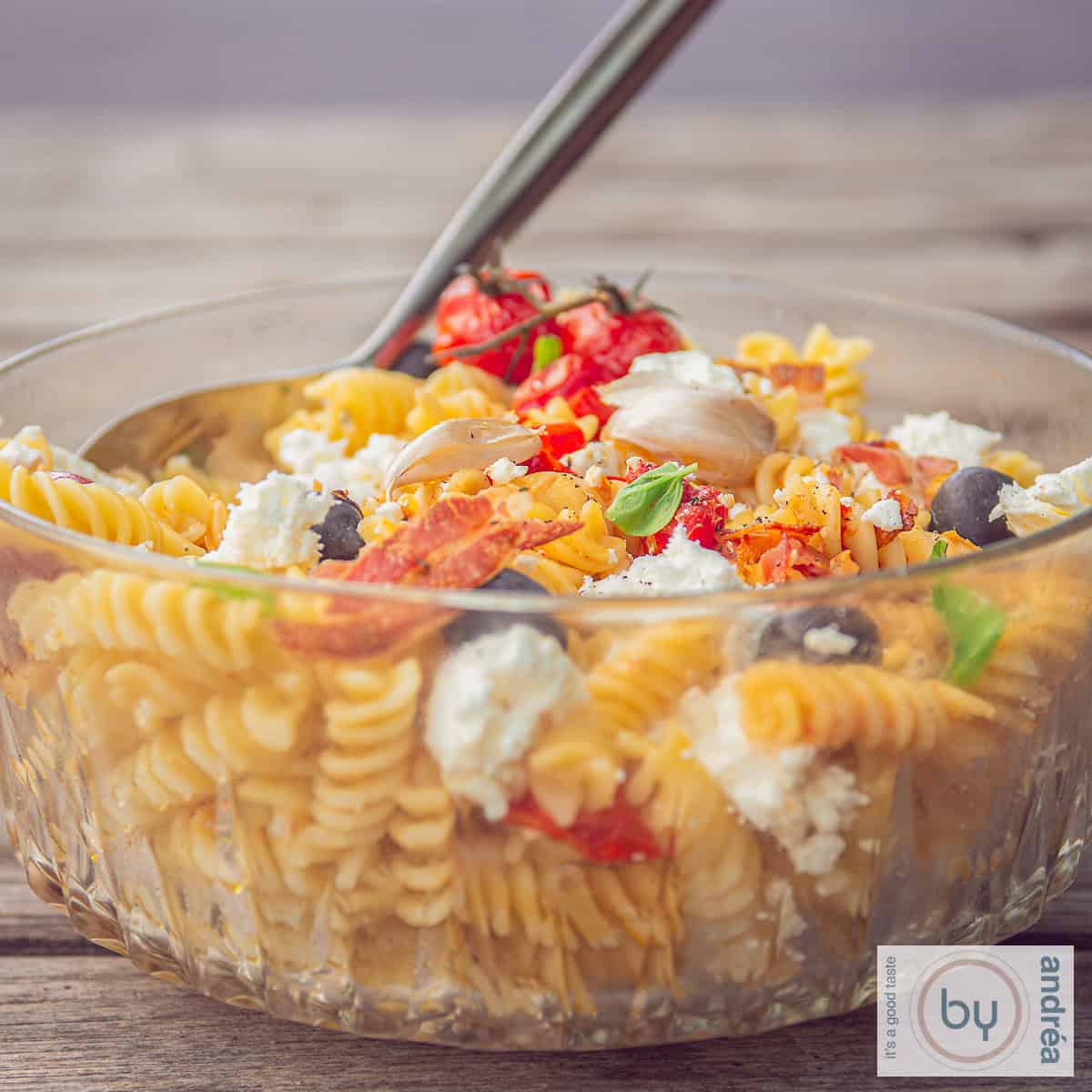 A square photo with a glass bowl on a wooden background filled with pasta salad, tomatoes, bacon and feta