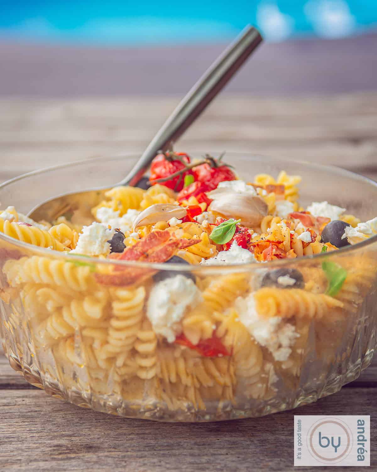 A photo of a glass bowl filled with a summer pasta salad with tomatoes, bacon and feta
