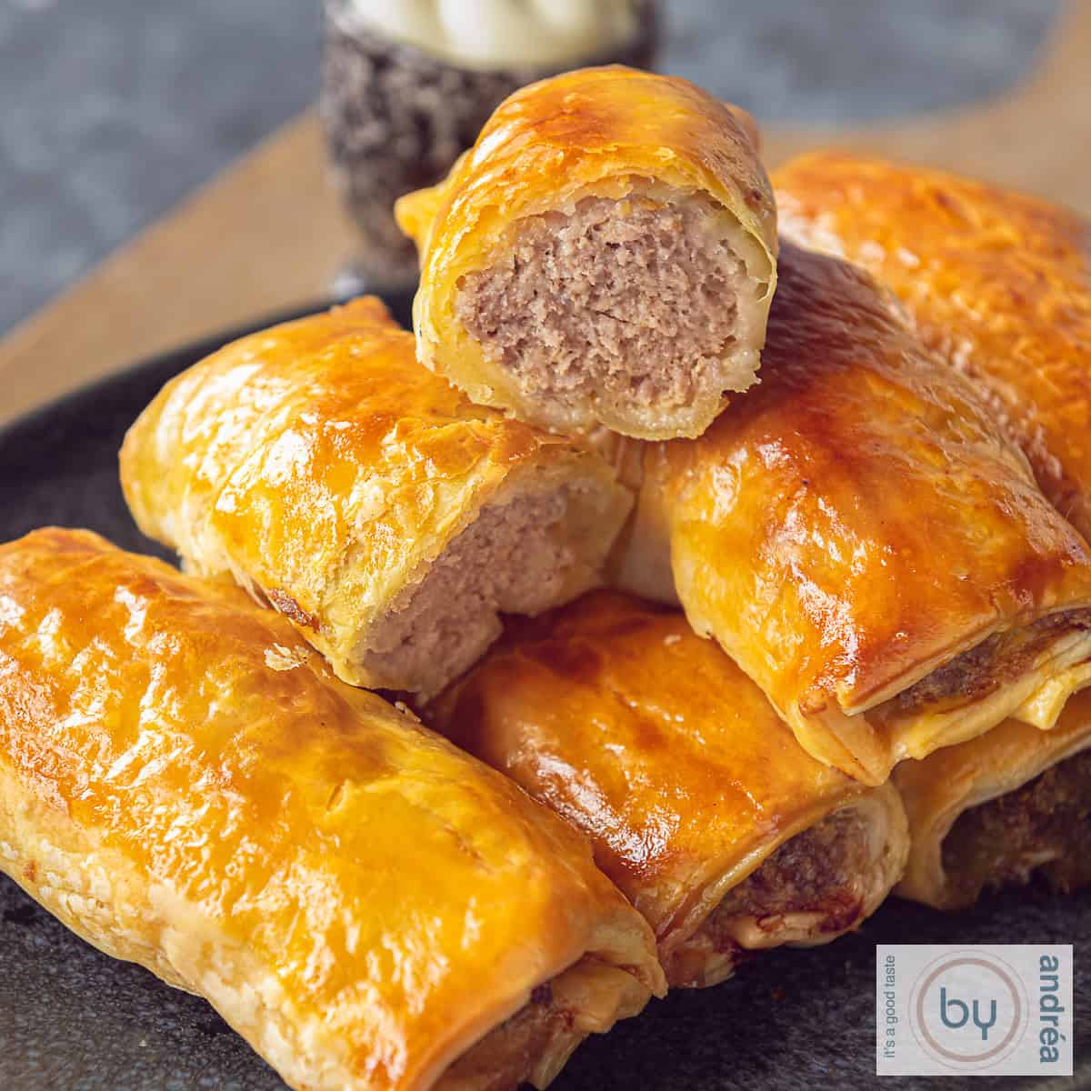 A square photo of a gray bowl with a stack of puff pastry sausage rolls. One roll cut in half