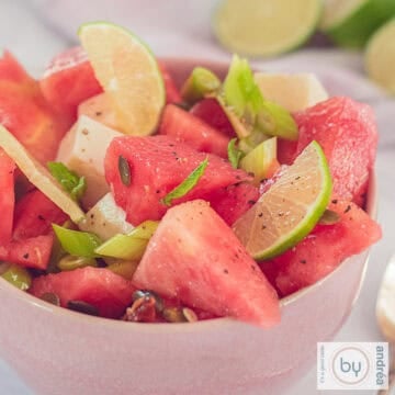 A square photo of a melon salad with goat cheese and nuts in a pink bowl.