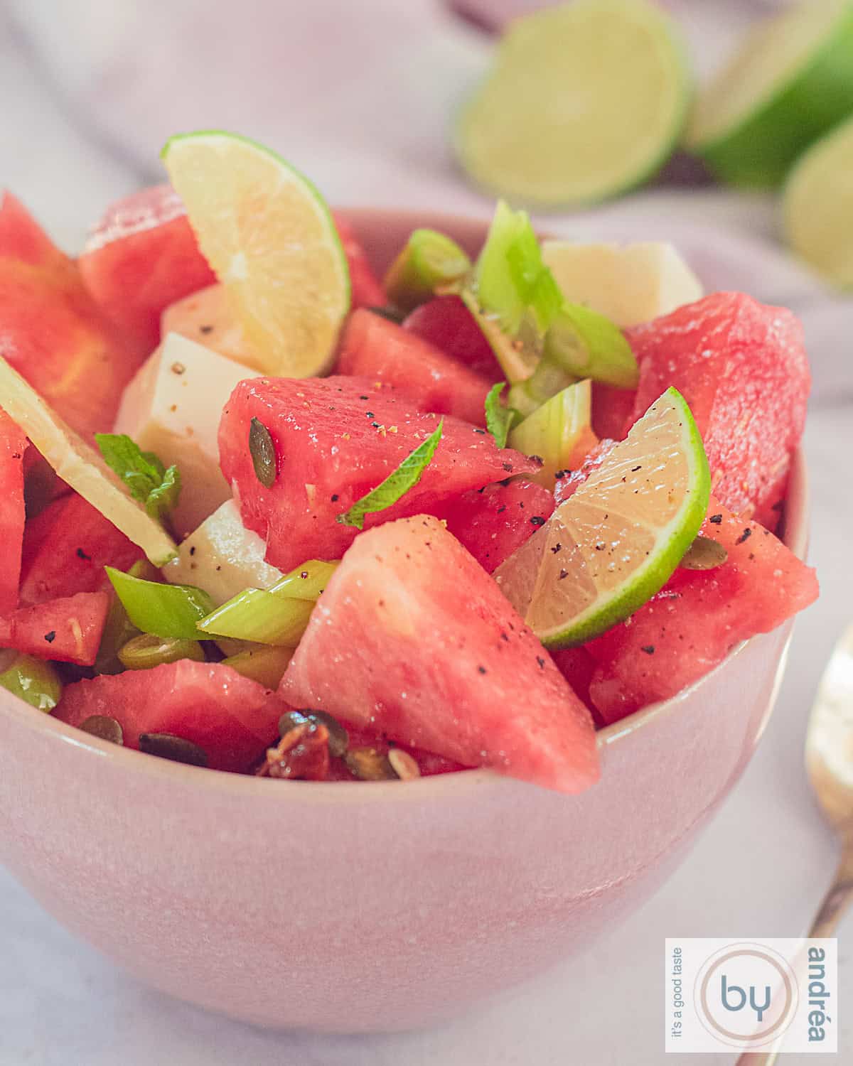 A photo of a pink bowl with watermelon, lime, spring onion, nuts and goat cheese.