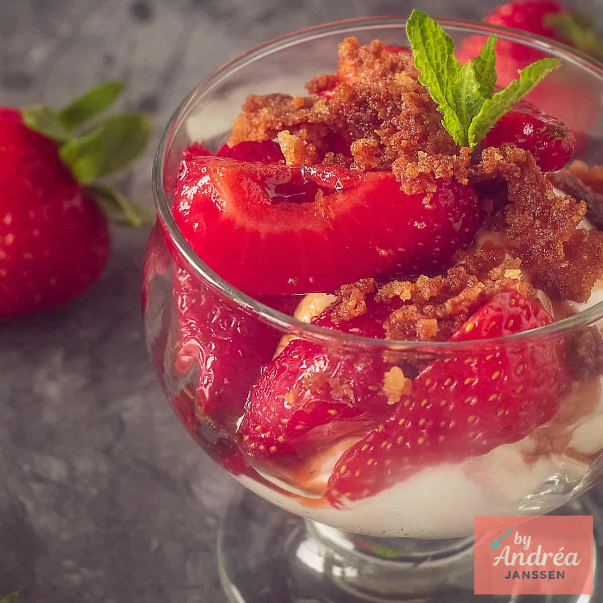 A square photo of a glass cup with yoghurt, marinated strawberries and vanilla crumbs on a dark gray background.