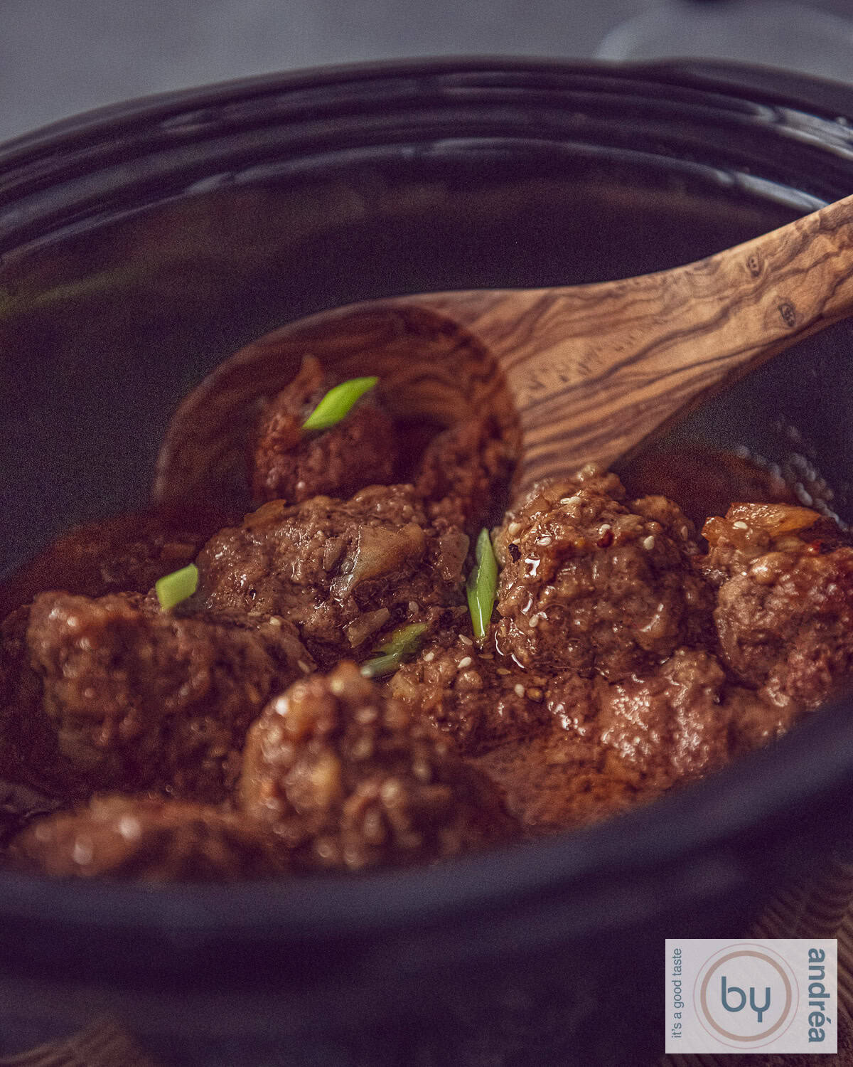 A slow cooker with meatballs in a sweet ketjap sauce and a wooden spoon in it.