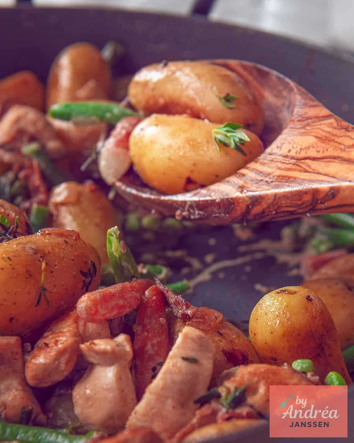 A portion of a skillet containing fried baby potatoes, chicken, and vegetables. A wooden spoon scoops out a scoop.