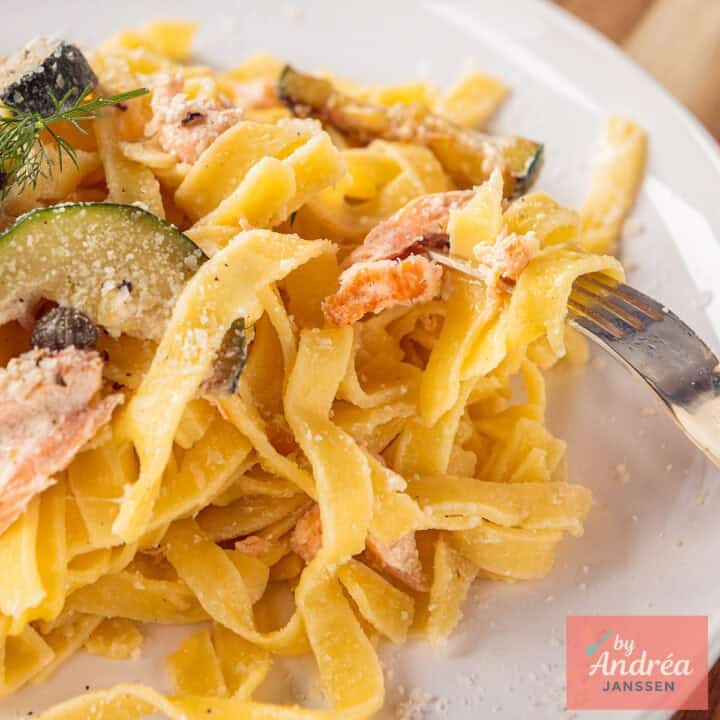 A square photo showing part of a white plate with pasta, zucchini, salmon, capers and creamy sauce.