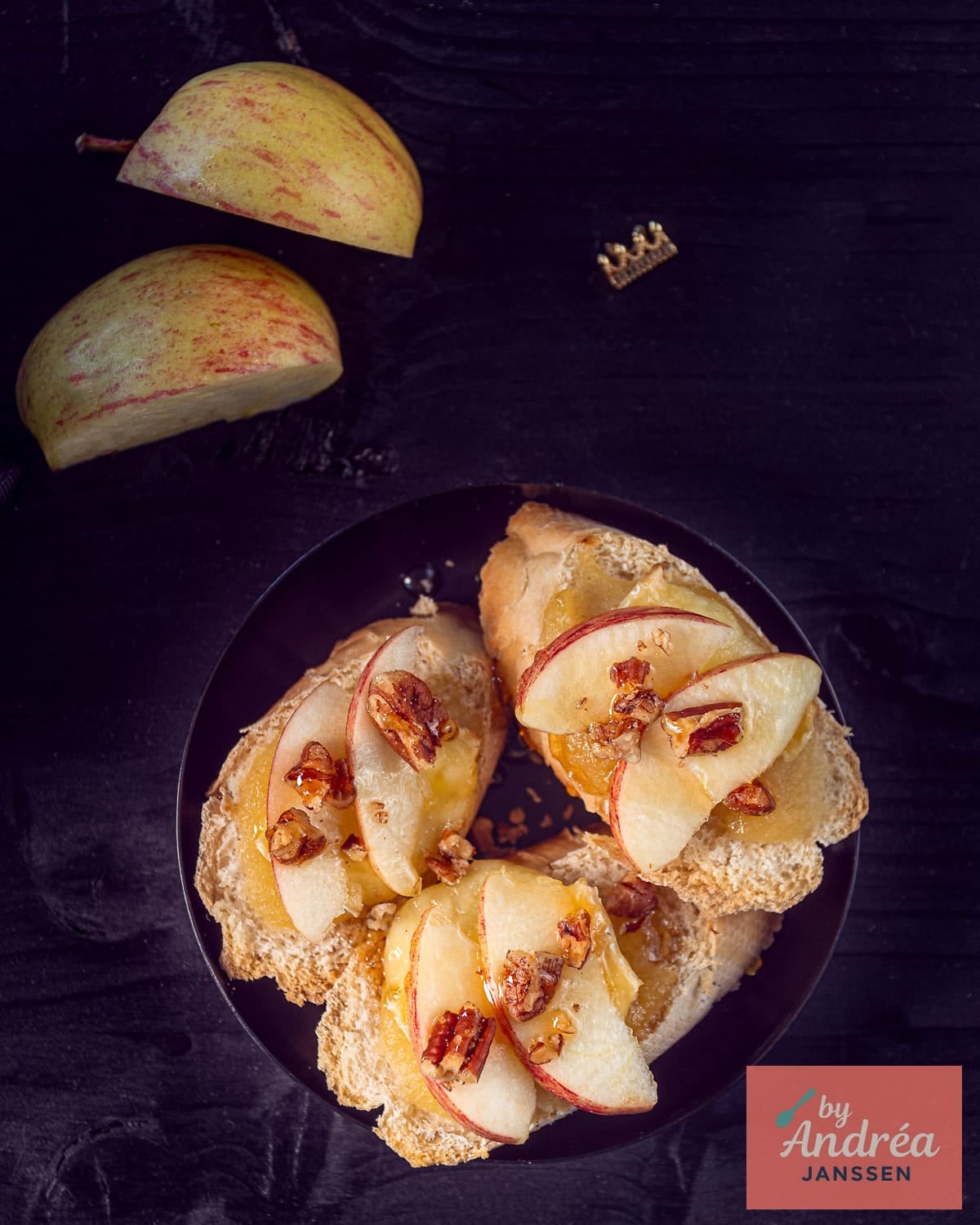 An overhead photo of three slices of baguette with apple, walnut, brie and honey on a dark background.