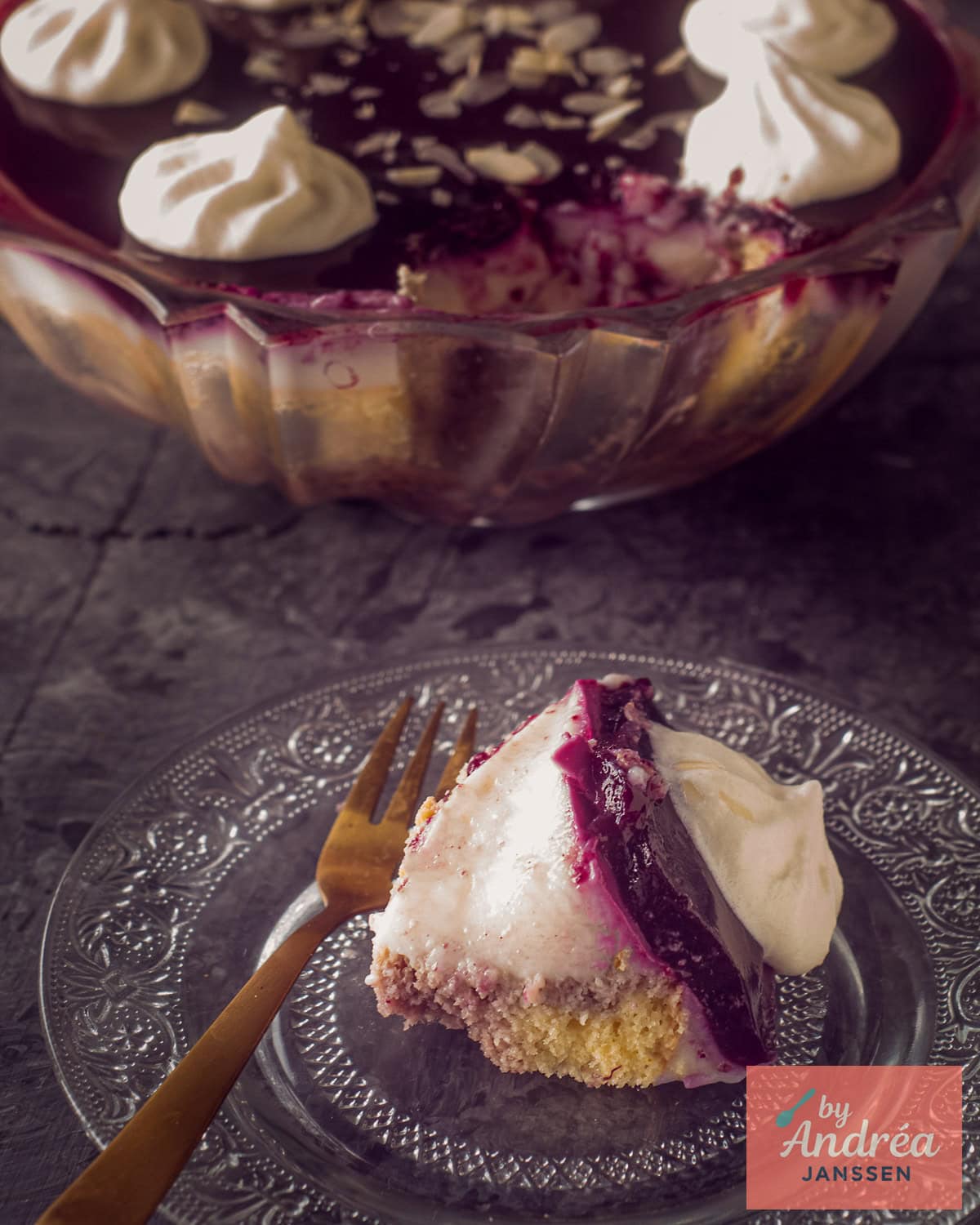 A piece of trifle with sherry, red fruit, and custard on a glass plate. The rest of the trifle is in the background.
