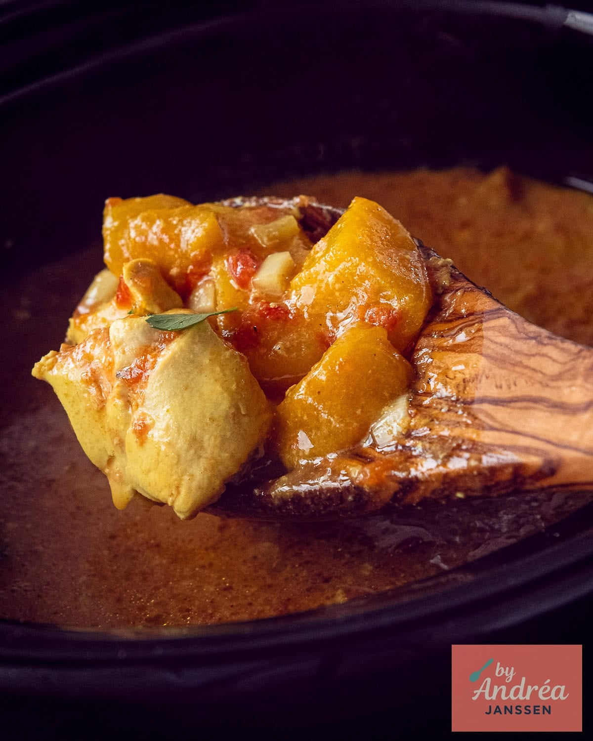 A slow cooker with a wooden spoon filled with chicken, curry, and mango.