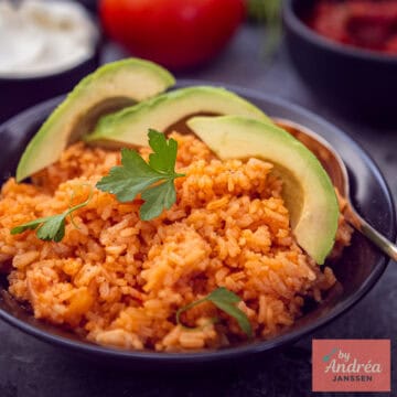 A square photo with a black bowl of red Mexican rice with avocado and parsley