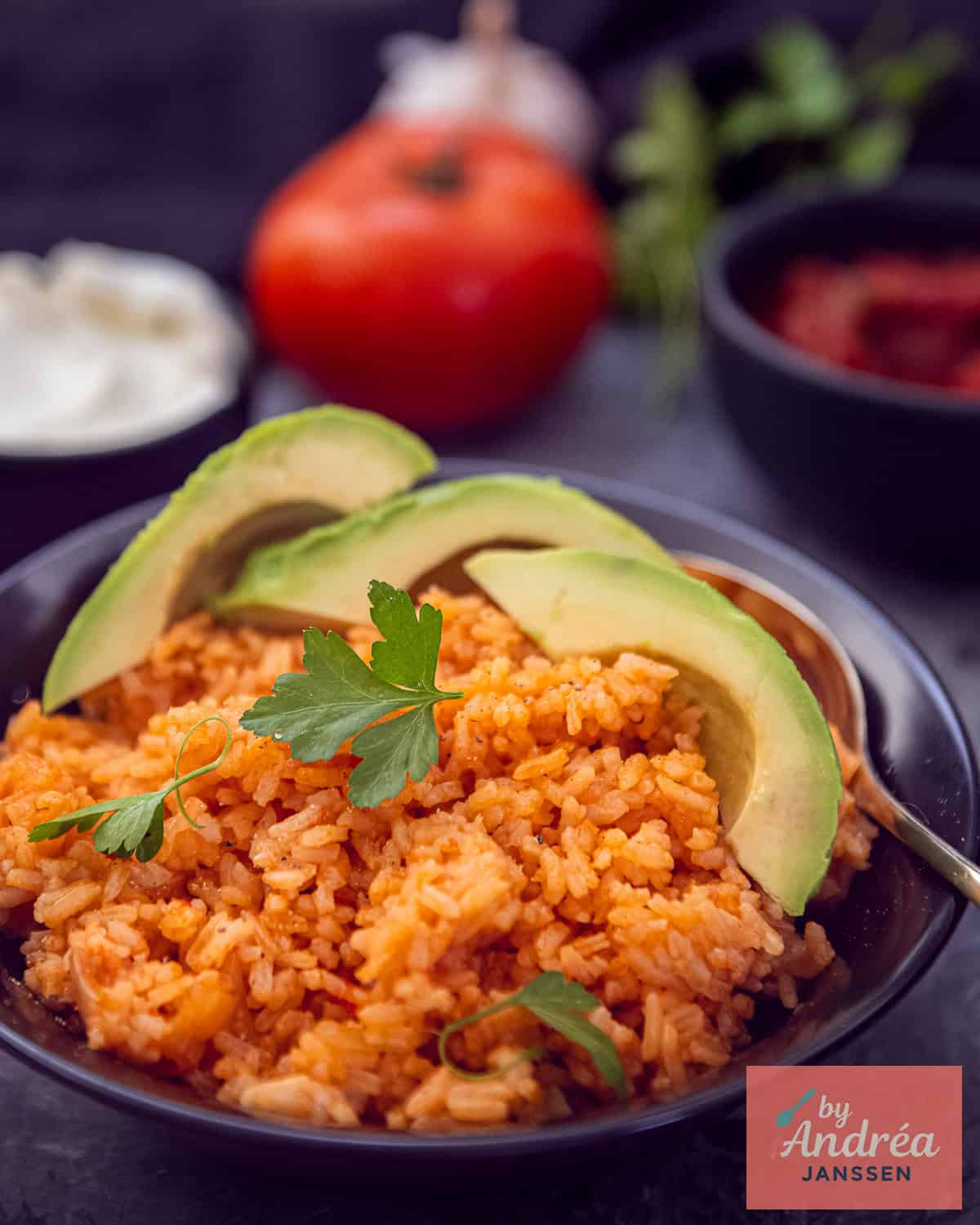 Een zwart schaaltje met Mexicaanse rijst met peterselie en avocado. In de achtergrond, tomaat, peterselie, salsa en creme fraiche.