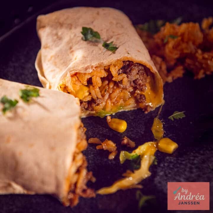 A square photo of a cut-open burrito with a filling of rice, corn, ground beef, cheese, cabbage, tomato, and onion.