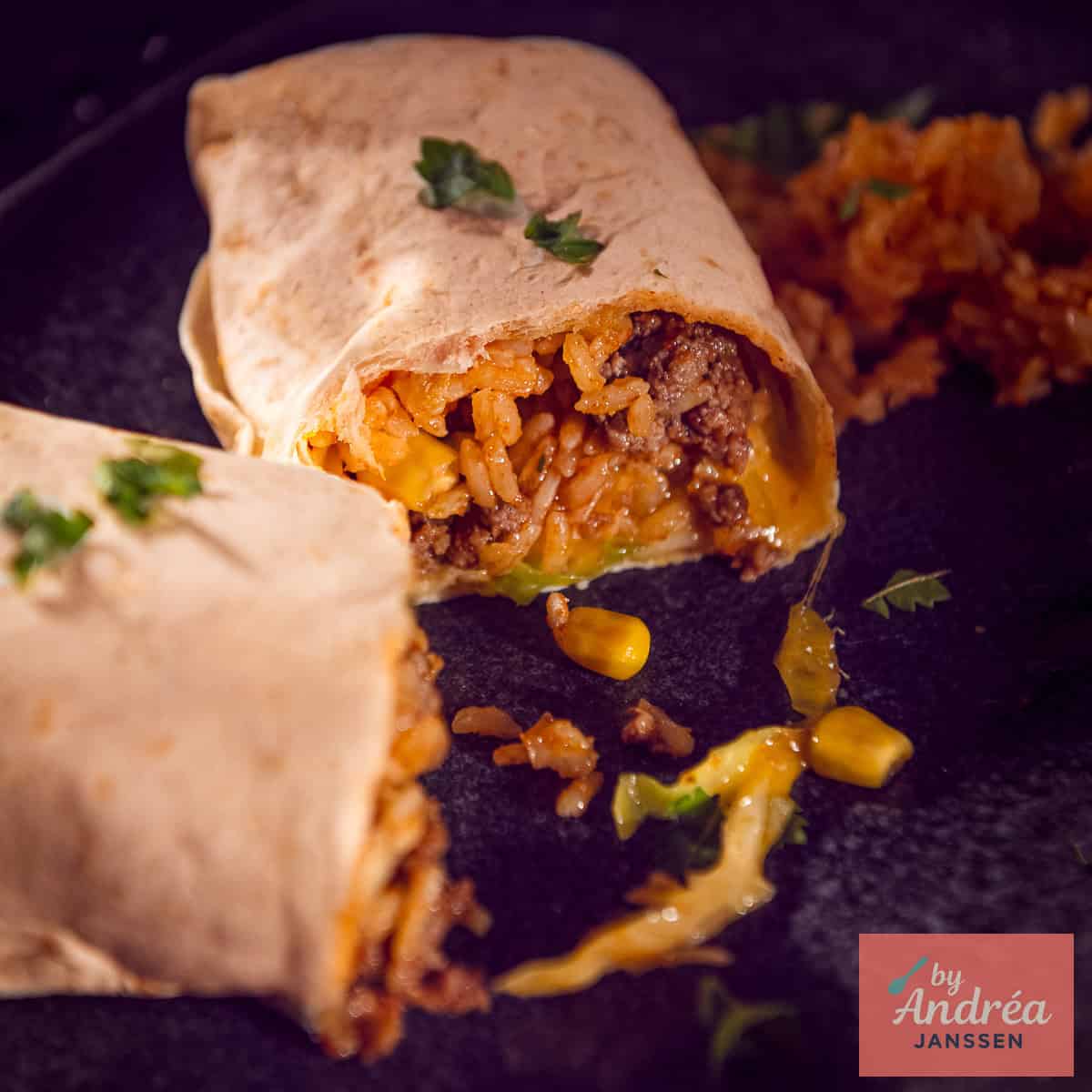 A square photo of a cut-open burrito with a filling of rice, corn, ground beef, cheese, cabbage, tomato, and onion.