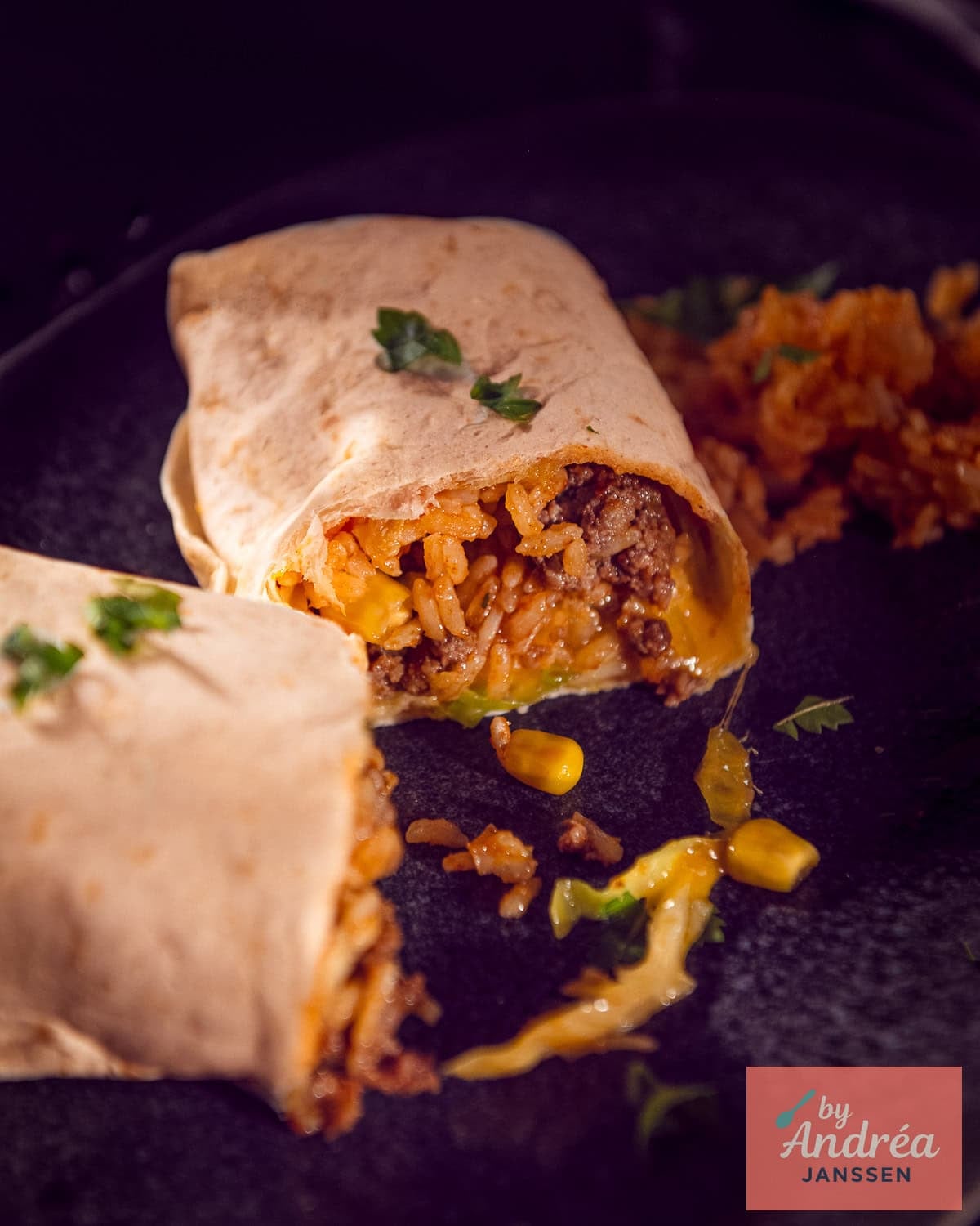A grey plate with a burrito cut open so you can clearly see the filling with rice, ground beef, corn, cabbage, tomato, and cheese.