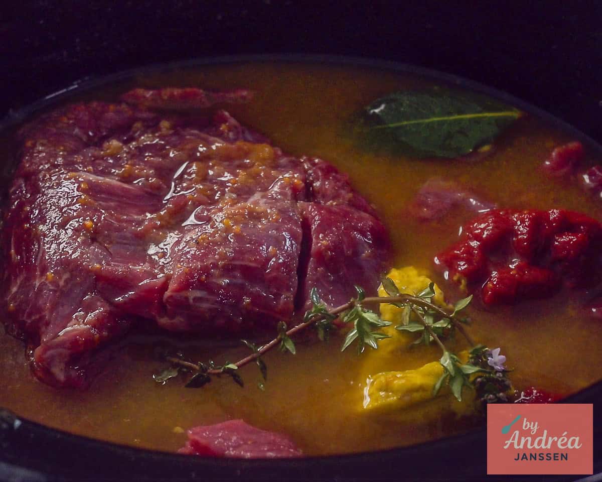A slow cooker filled with beef, broth, mustard, tomato paste, herbs, and other seasonings