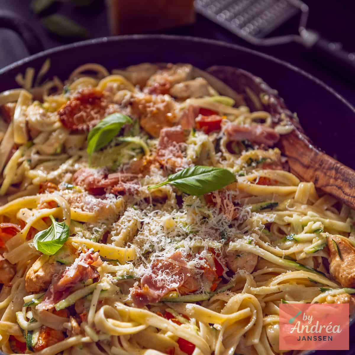 A square photo of a sauté pan with pasta, chicken, and creamy sauce with bacon.