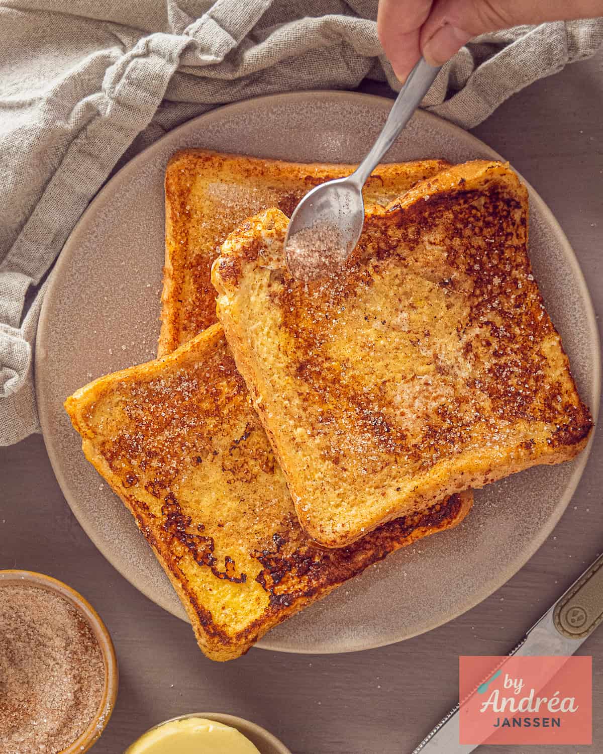 A beige plate with three French toast slices in a stack. A spoon sprinkles some cinnamon sugar over them.