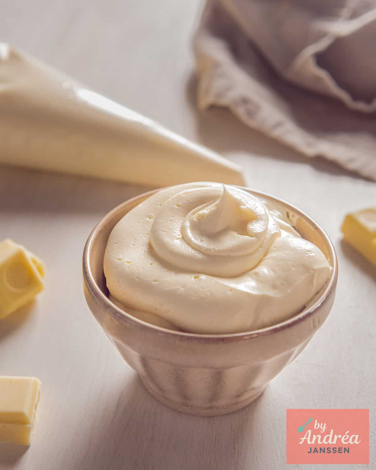 A beige bowl of Creamy White Chocolate Buttercream. In the background, a piping bag with more cream.