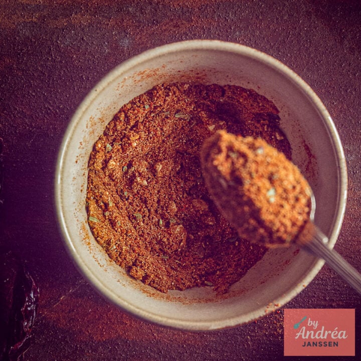 Overhead view of a bowl filled with reddish-brown chili con carne seasoning and a spoonful of the spice mix.