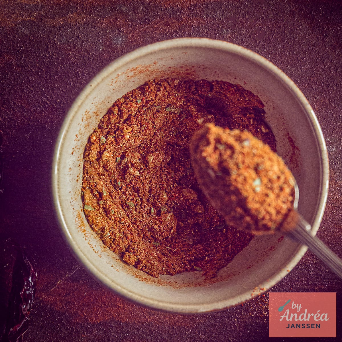 Overhead view of a bowl filled with reddish-brown chili con carne seasoning and a spoonful of the spice mix.