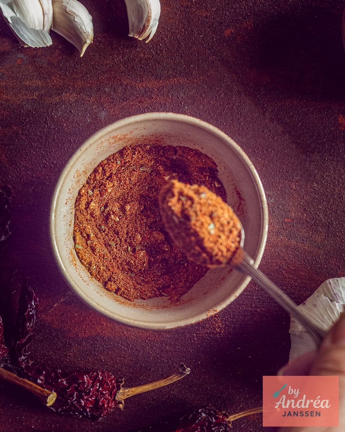 Overhead view of homemade chili con carne seasoning in a bowl with a full spoon, garlic cloves, and dried chili peppers.