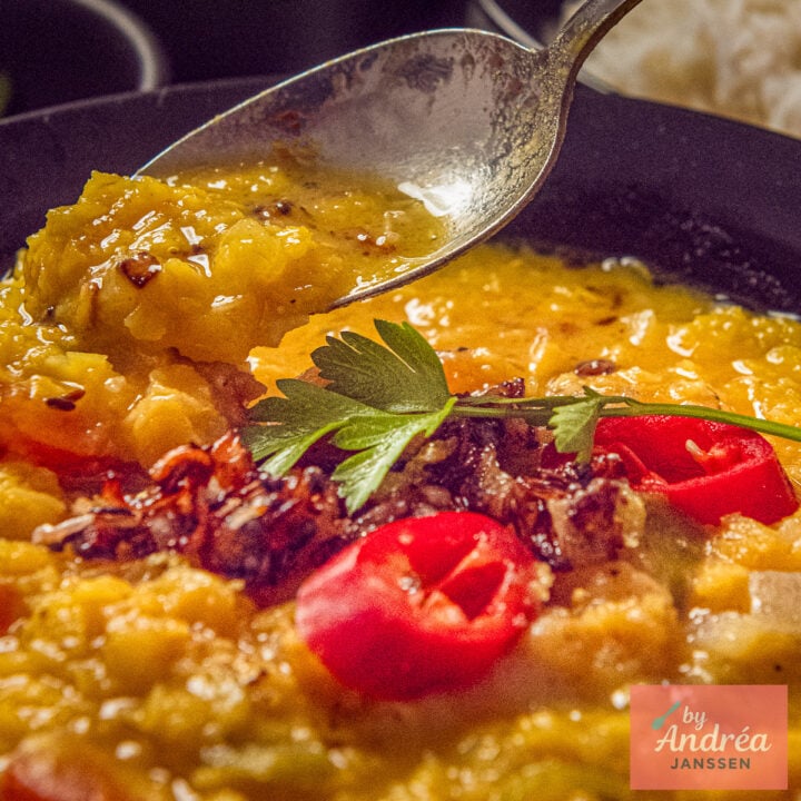 A spoonful of yellow split pea dal above a bowl topped with tadka, sliced red chili, and fresh herbs.