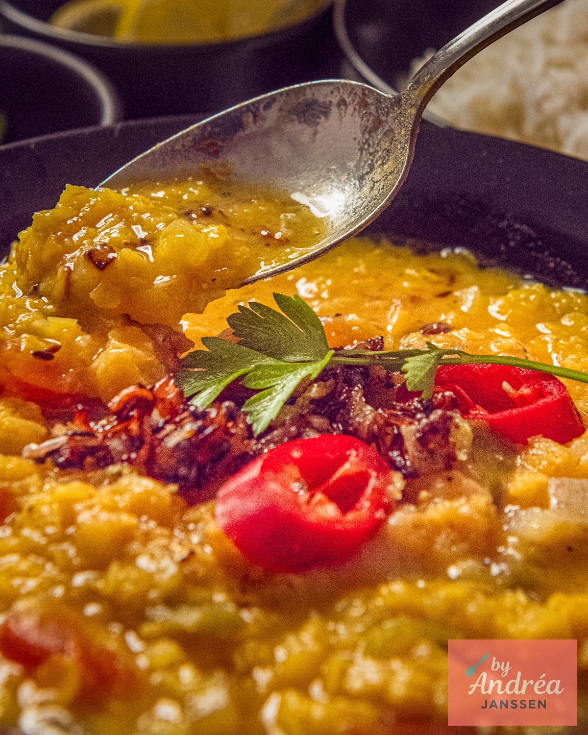 A spoonful of yellow split pea dal above a bowl topped with tadka, sliced red chili, and fresh herbs.