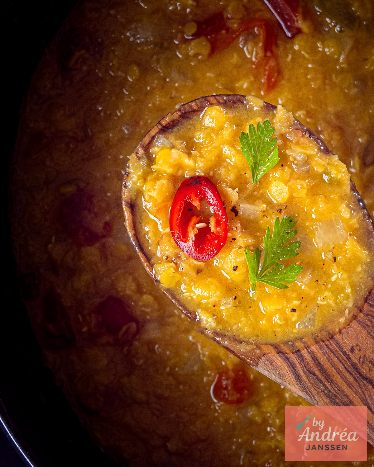 Wooden spoon holding yellow split pea dal with a red chili slice and green herbs above the slow cooker.