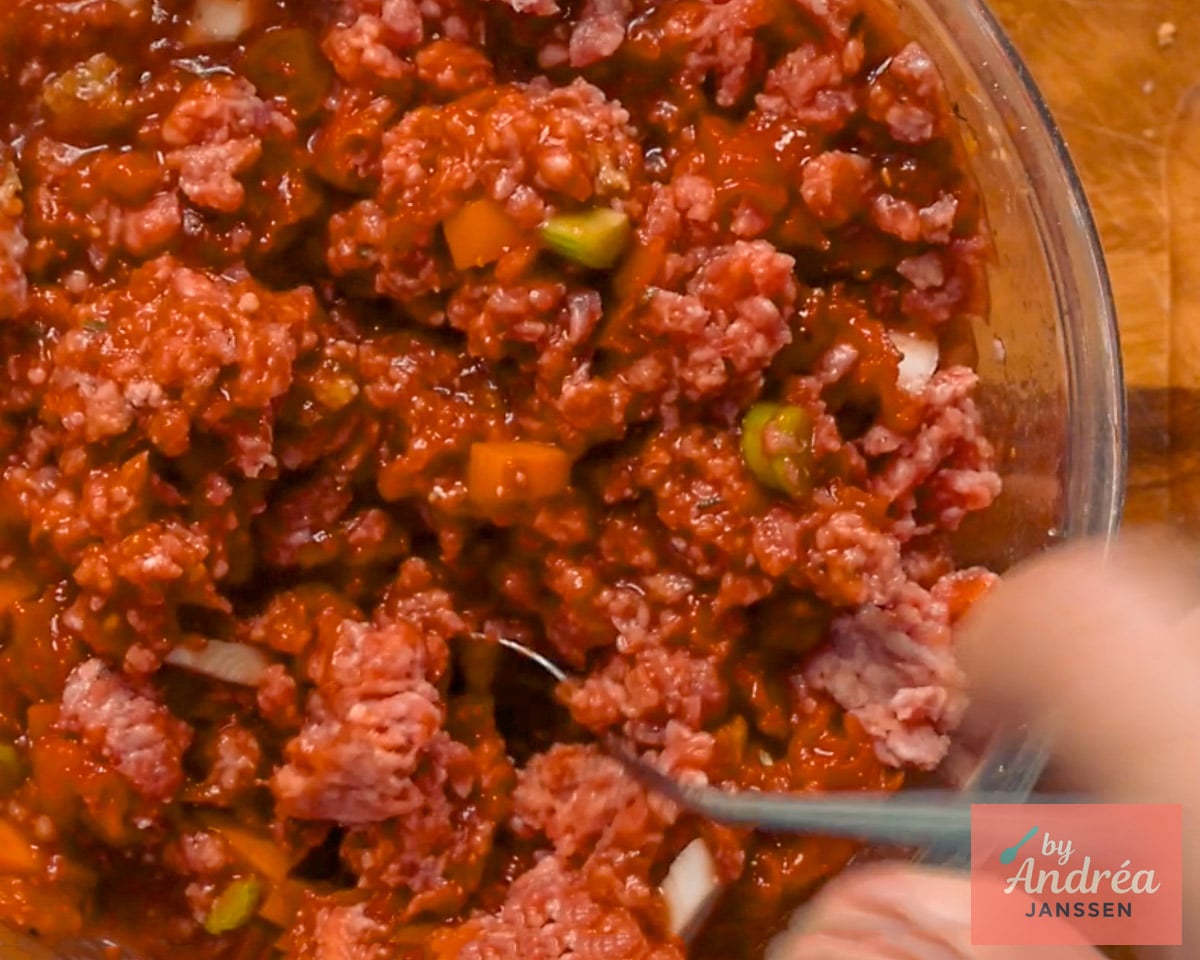 Ground beef being mixed with tomato sauce, carrot, and celery in a glass bowl.