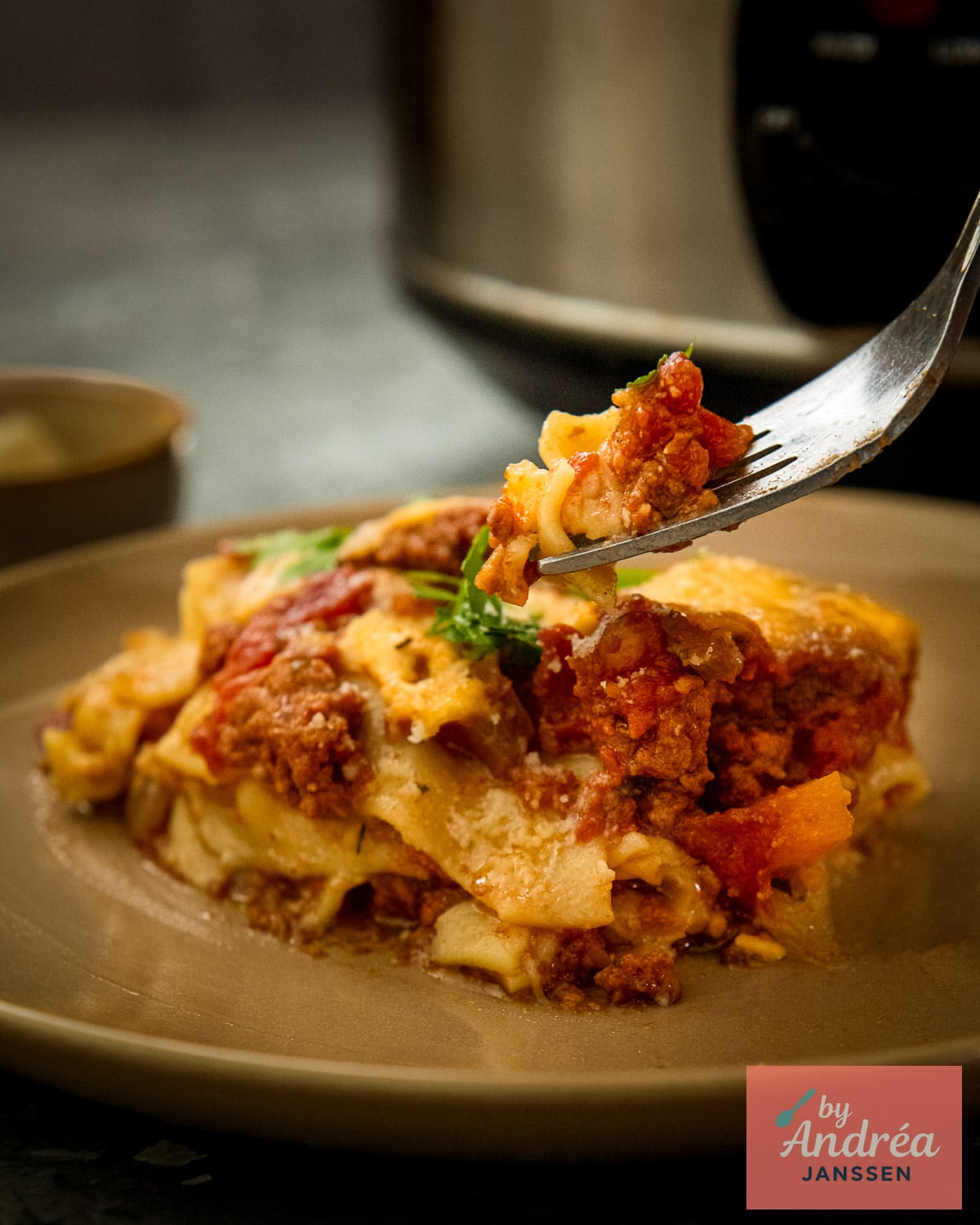A fork lifts a bite of lasagna with meat sauce and cheese above a plate, with the slow cooker in the background.
