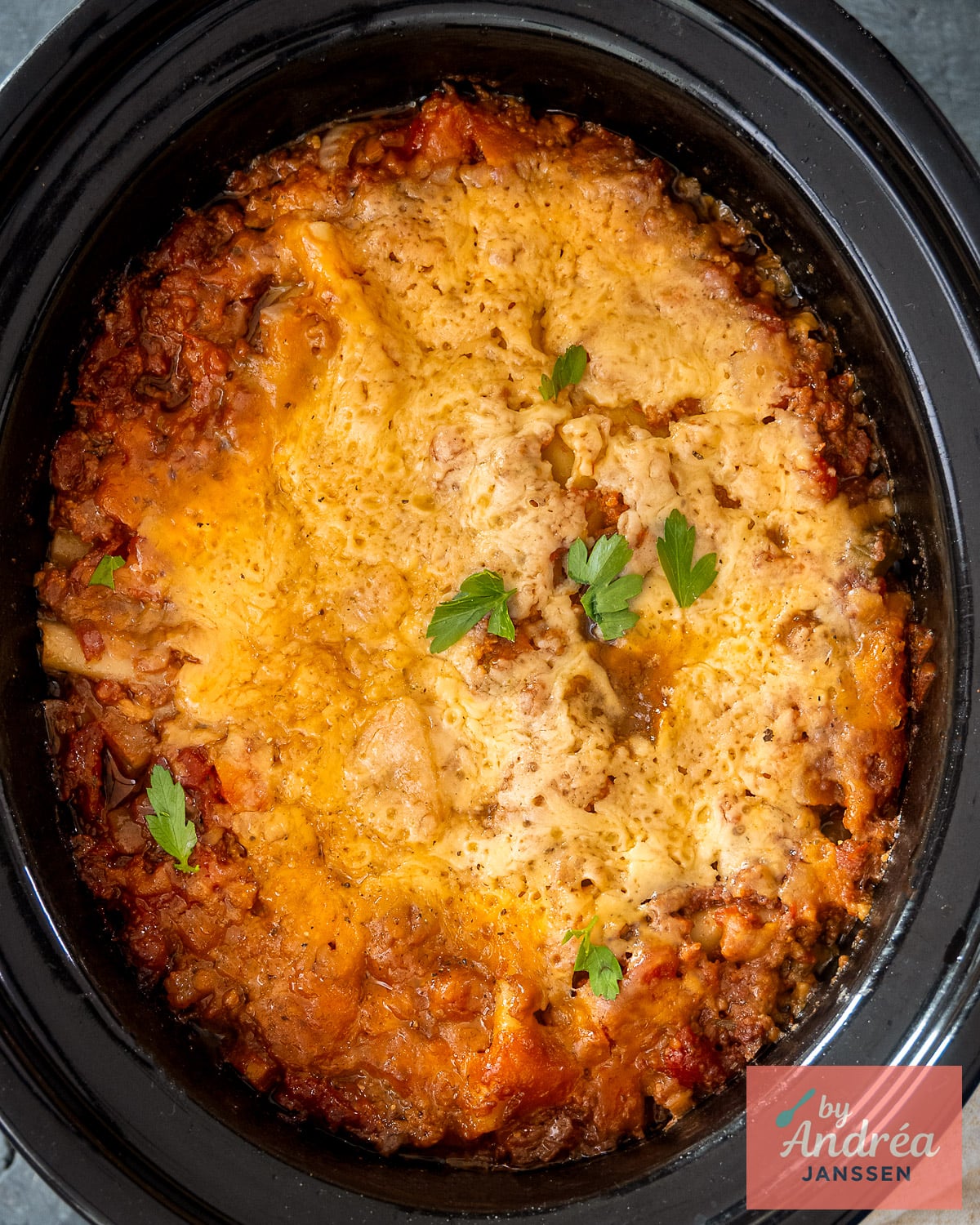 Overhead view of lasagna with a golden cheese topping and parsley in a black slow cooker.
