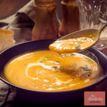 A square picture with A spoonful of creamy butternut squash soup above a black bowl topped with cream and pepitas, with bread alongside.