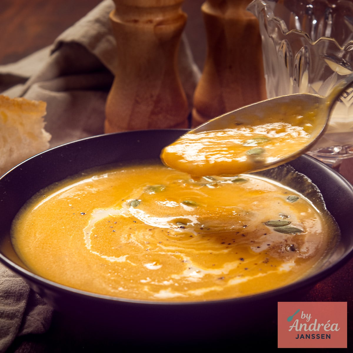 A square picture with A spoonful of creamy butternut squash soup above a black bowl topped with cream and pepitas, with bread alongside.