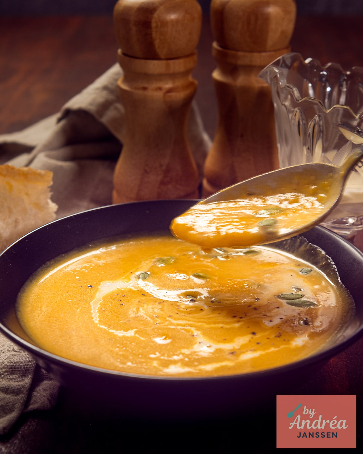 A spoonful of creamy butternut squash soup above a black bowl topped with cream and pepitas, with bread alongside.