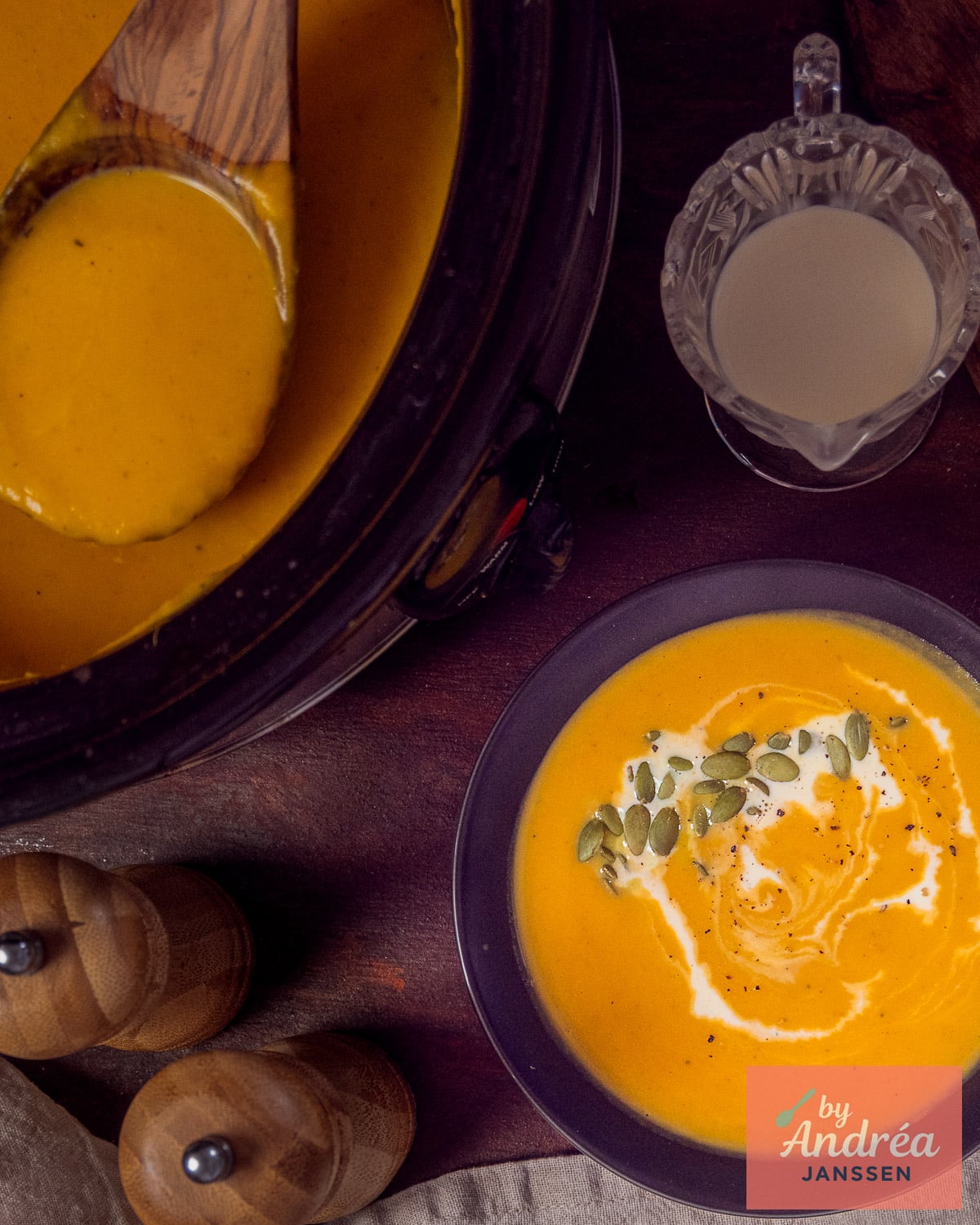 Overhead view of butternut squash soup topped with cream and pepitas beside the filled slow cooker with a wooden ladle.