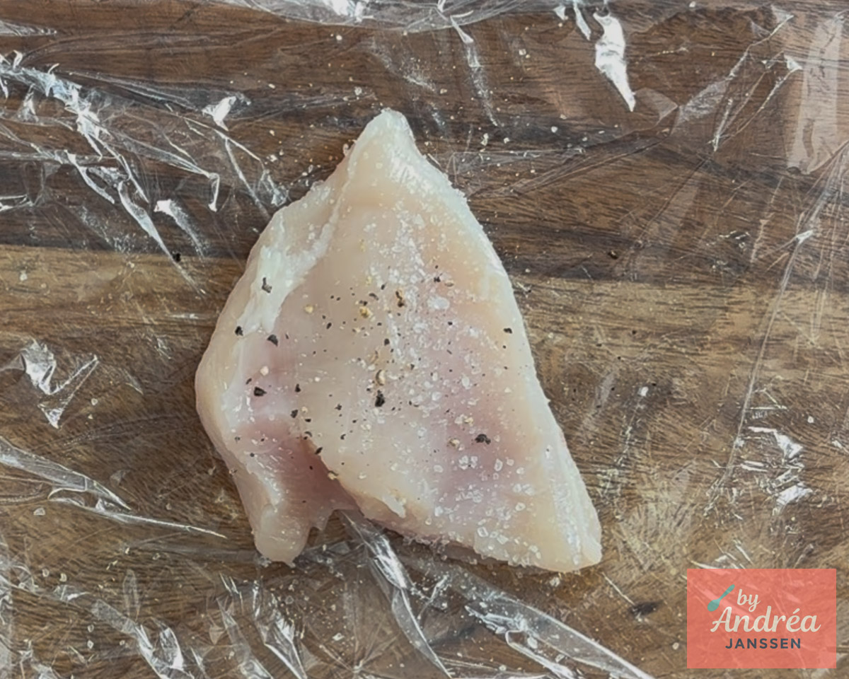 Raw chicken breast sprinkled with salt and pepper on plastic wrap on a wooden cutting board.
