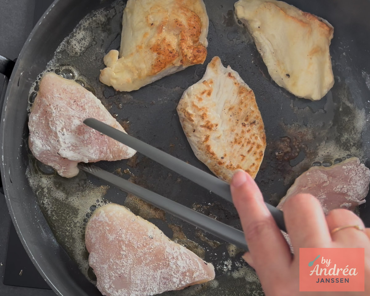 Flour-coated chicken breasts cooking in a skillet and being turned with kitchen tongs.
