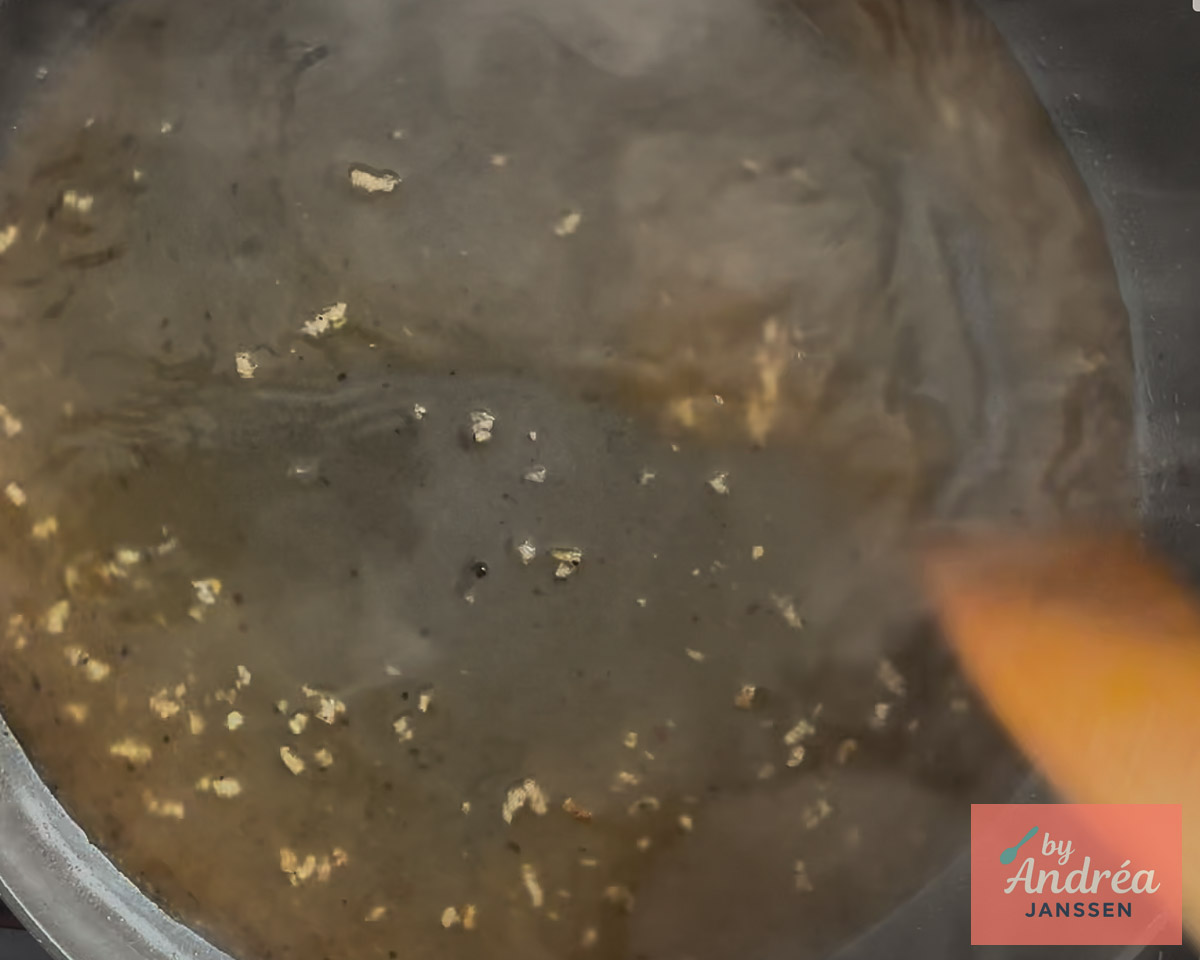 Honey lemon sauce with bits of garlic in a skillet, with a wooden spoon on the right.