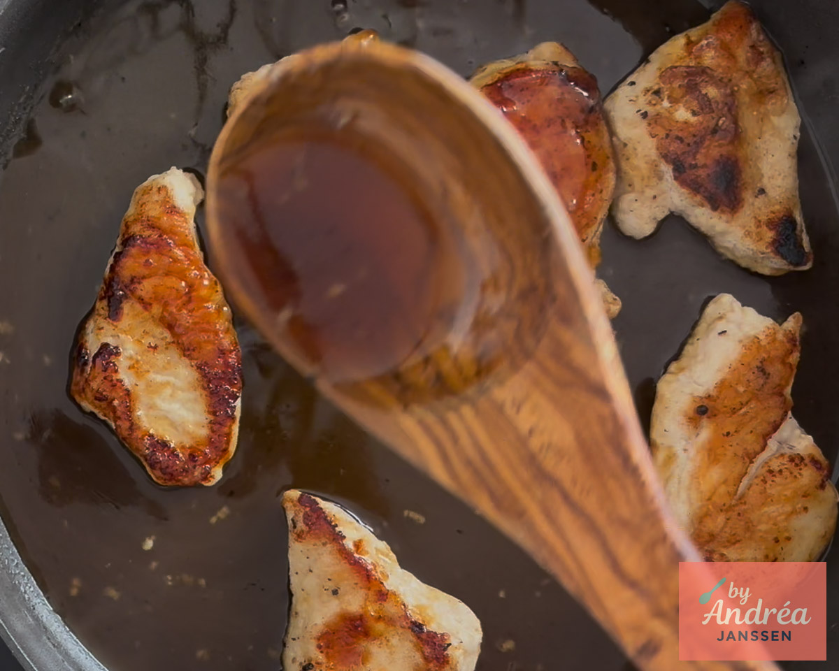 A wooden spoon filled with honey lemon sauce held over browned chicken breasts in a skillet.