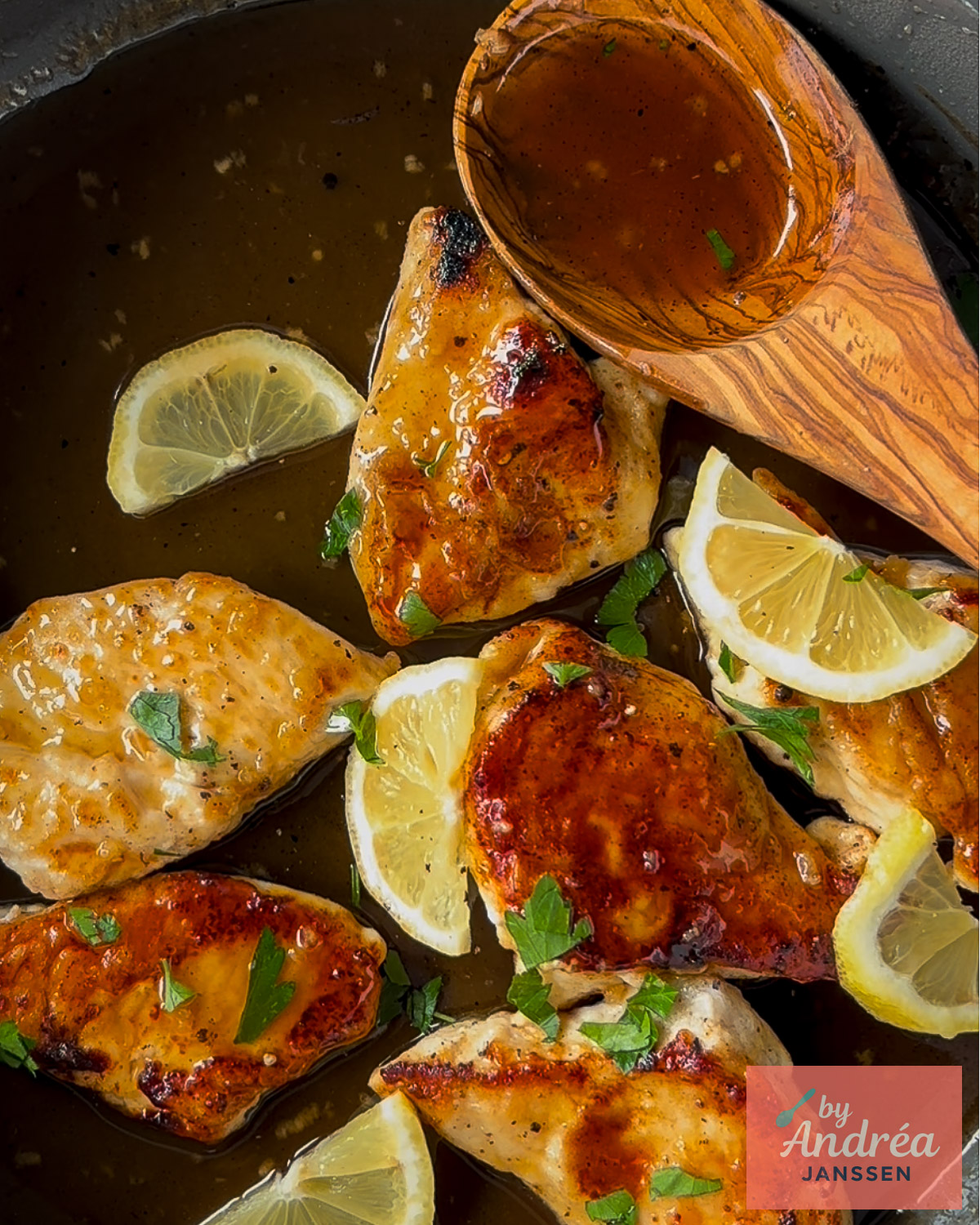 Overhead view of chicken breasts in honey lemon sauce with lemon slices, parsley, and a wooden spoon in the skillet.