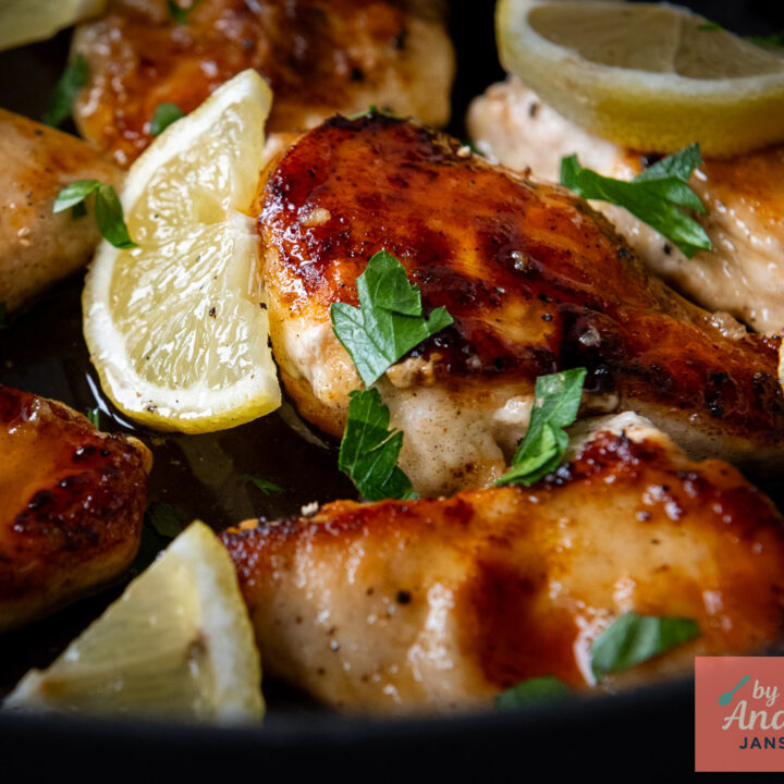 Golden-brown chicken breasts in honey lemon sauce with lemon slices and chopped parsley in a black skillet.