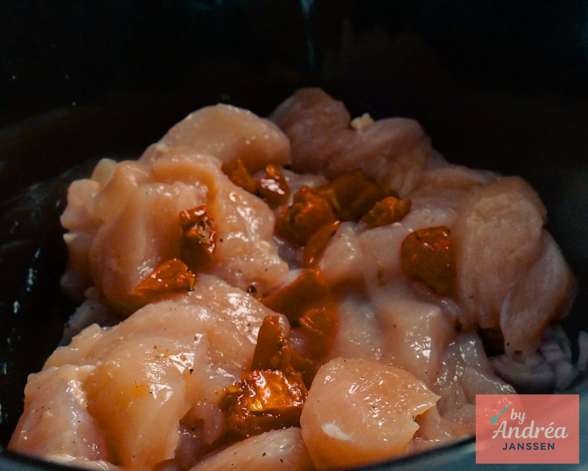 Raw chicken pieces and chopped sun-dried tomatoes layered over chopped shallot and garlic in the slow cooker.