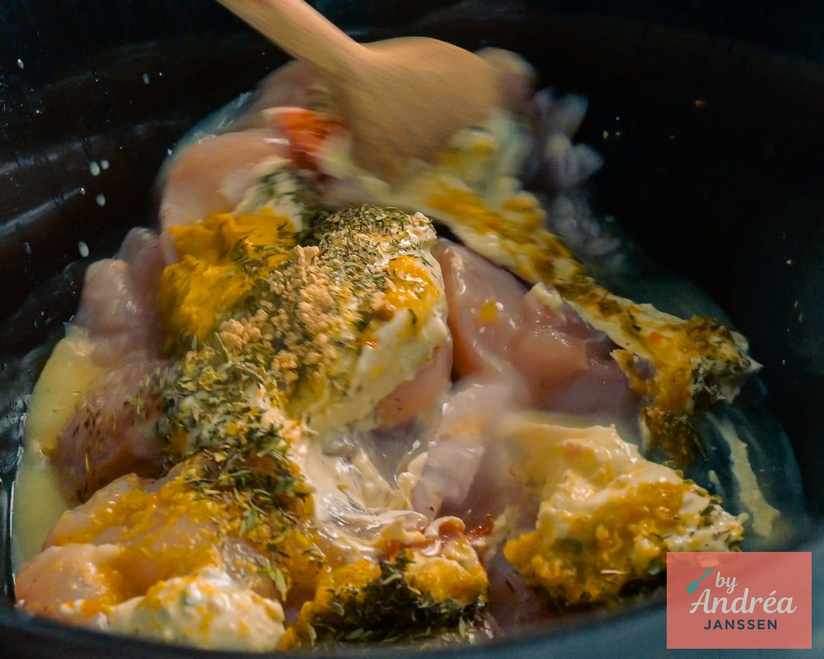 A wooden spoon mixes raw chicken with Boursin, Dijon mustard, dried herbs, and liquid in the slow cooker.