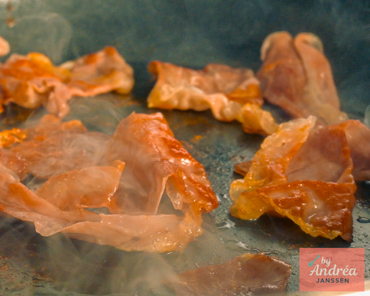 Thin slices of prosciutto cooking in a skillet, with golden brown, curled edges.