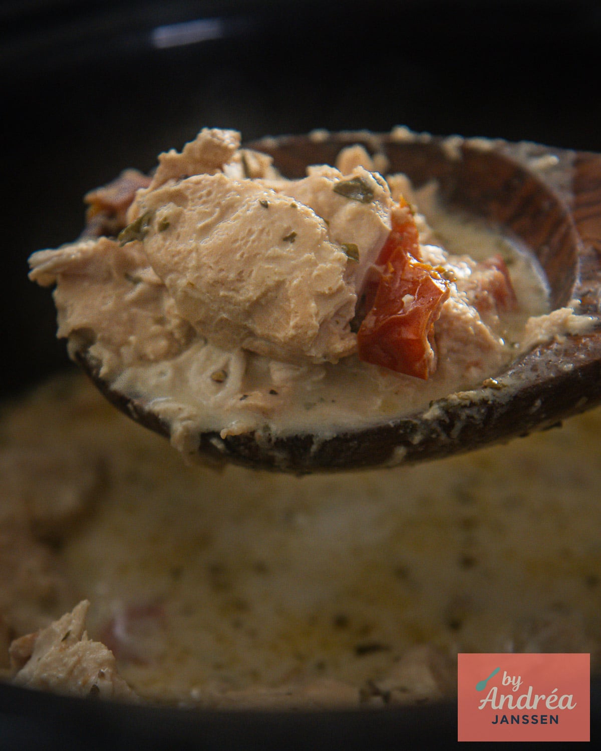 Wooden spoon holding chicken and sun-dried tomato pieces coated in creamy Boursin sauce above the slow cooker.