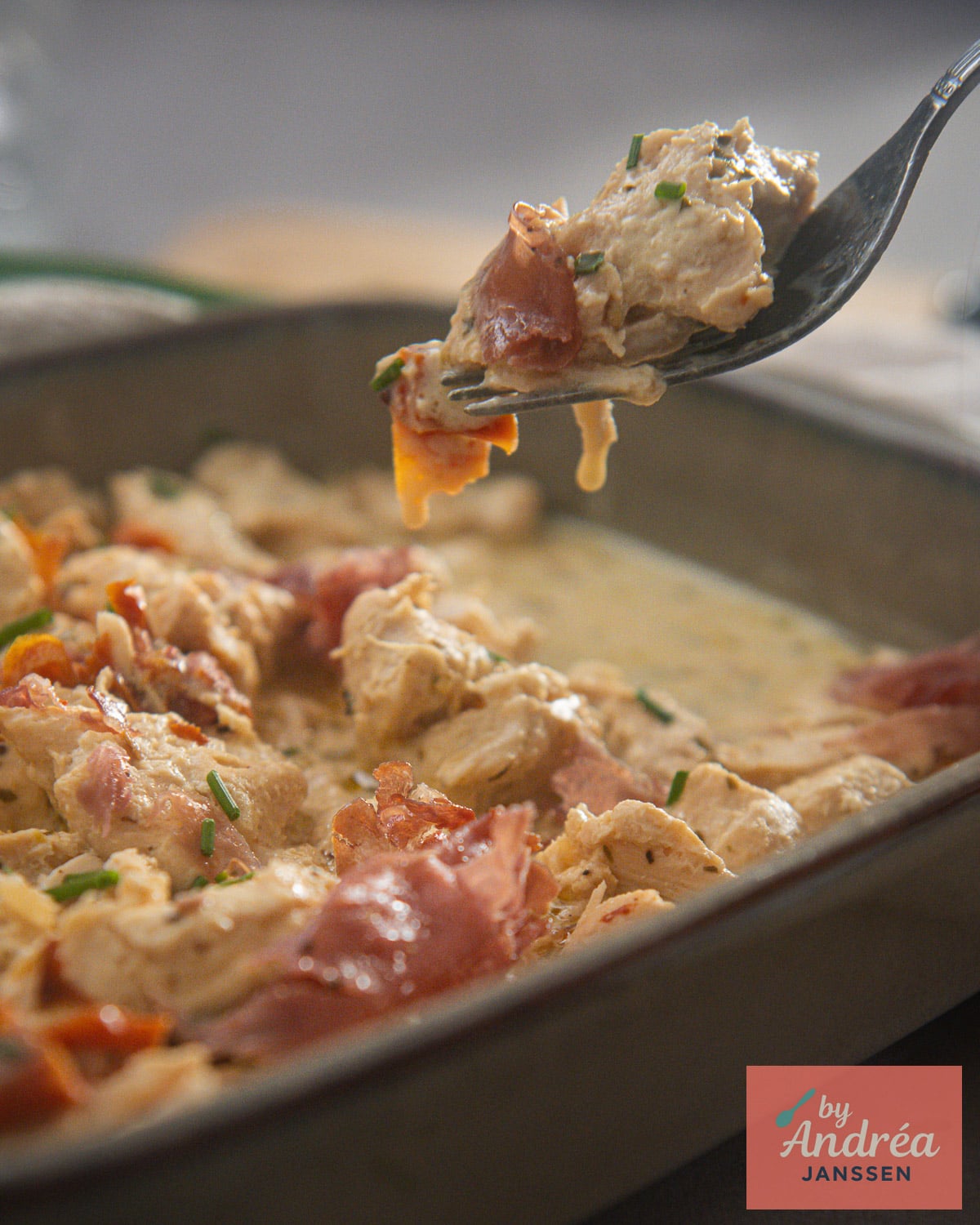Fork holding a bite of chicken in Boursin sauce with crispy prosciutto and chives above the serving dish.