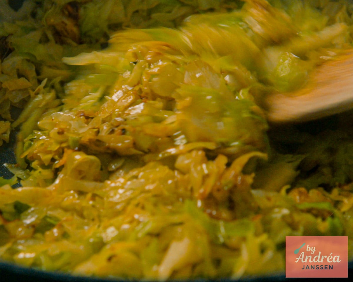 Shredded pointed cabbage and onion coated in curry powder being stirred with a wooden spoon in a skillet.