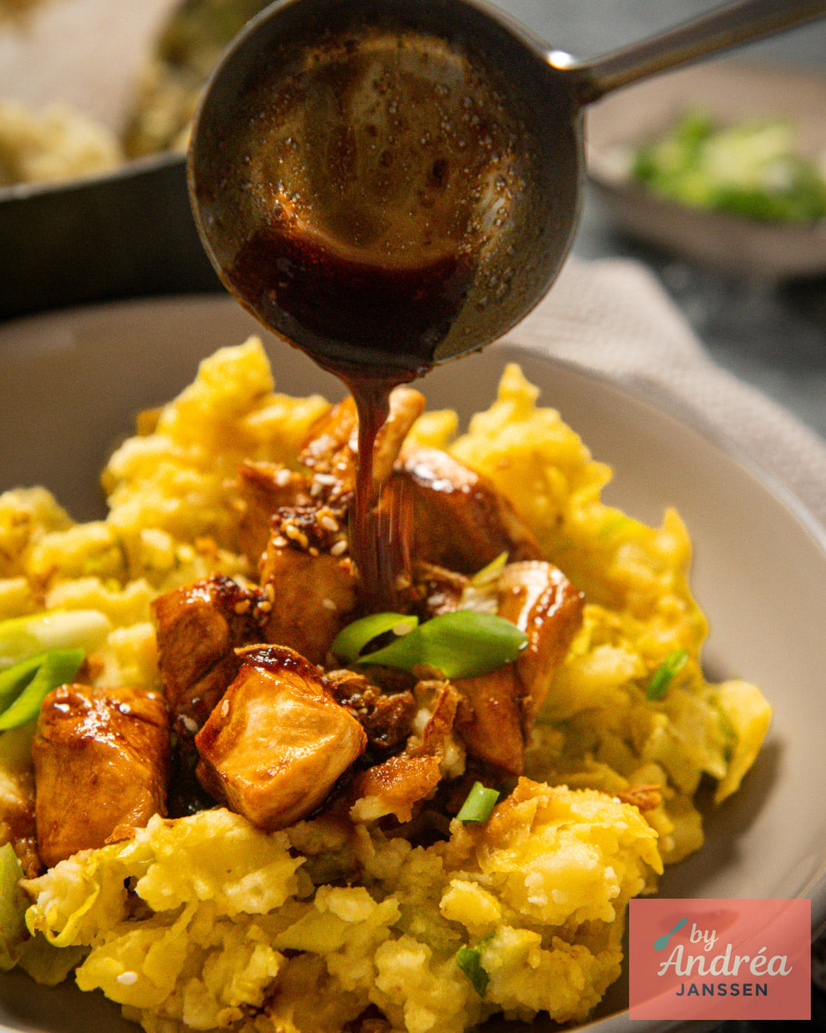 Dark sweet soy sauce pouring from a spoon over chicken on curried cabbage mash with green onions and sesame seeds.